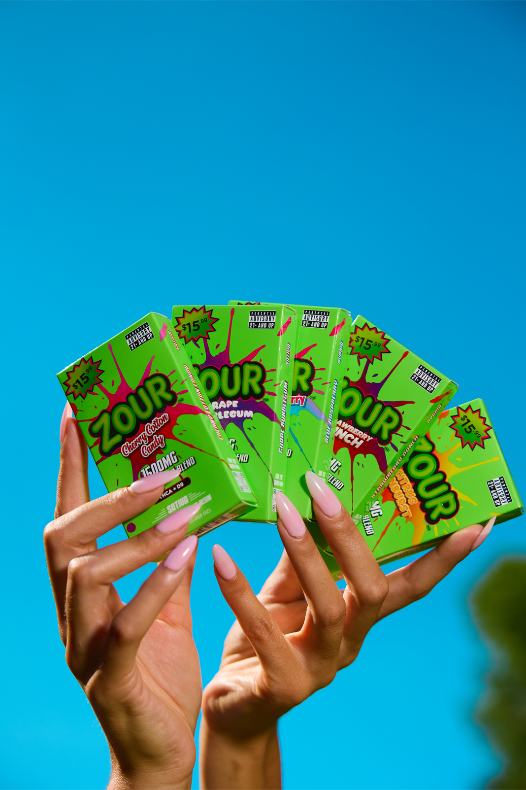 Two hands holding a fan of Sour Patch Kids candy boxes against a bright blue sky background.
