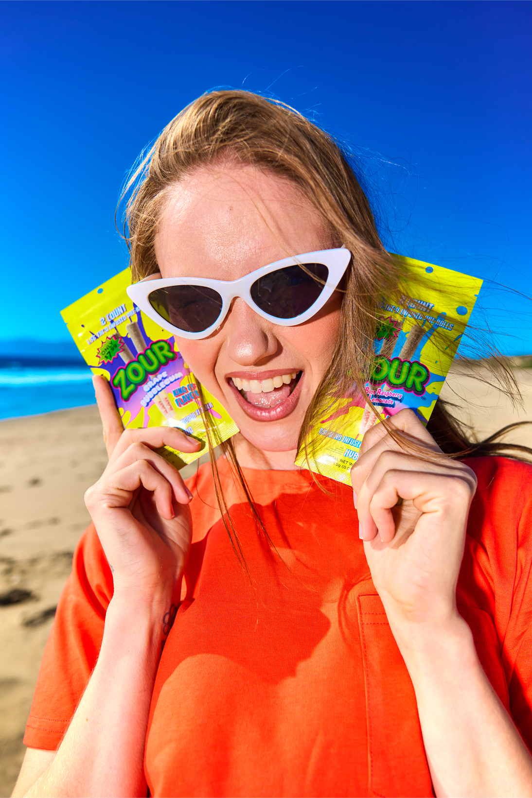 A woman on a beach wearing white cat-eye sunglasses and an orange top holds two bright yellow Zour Preroll 2 counts near her face against a blue sky and ocean background.