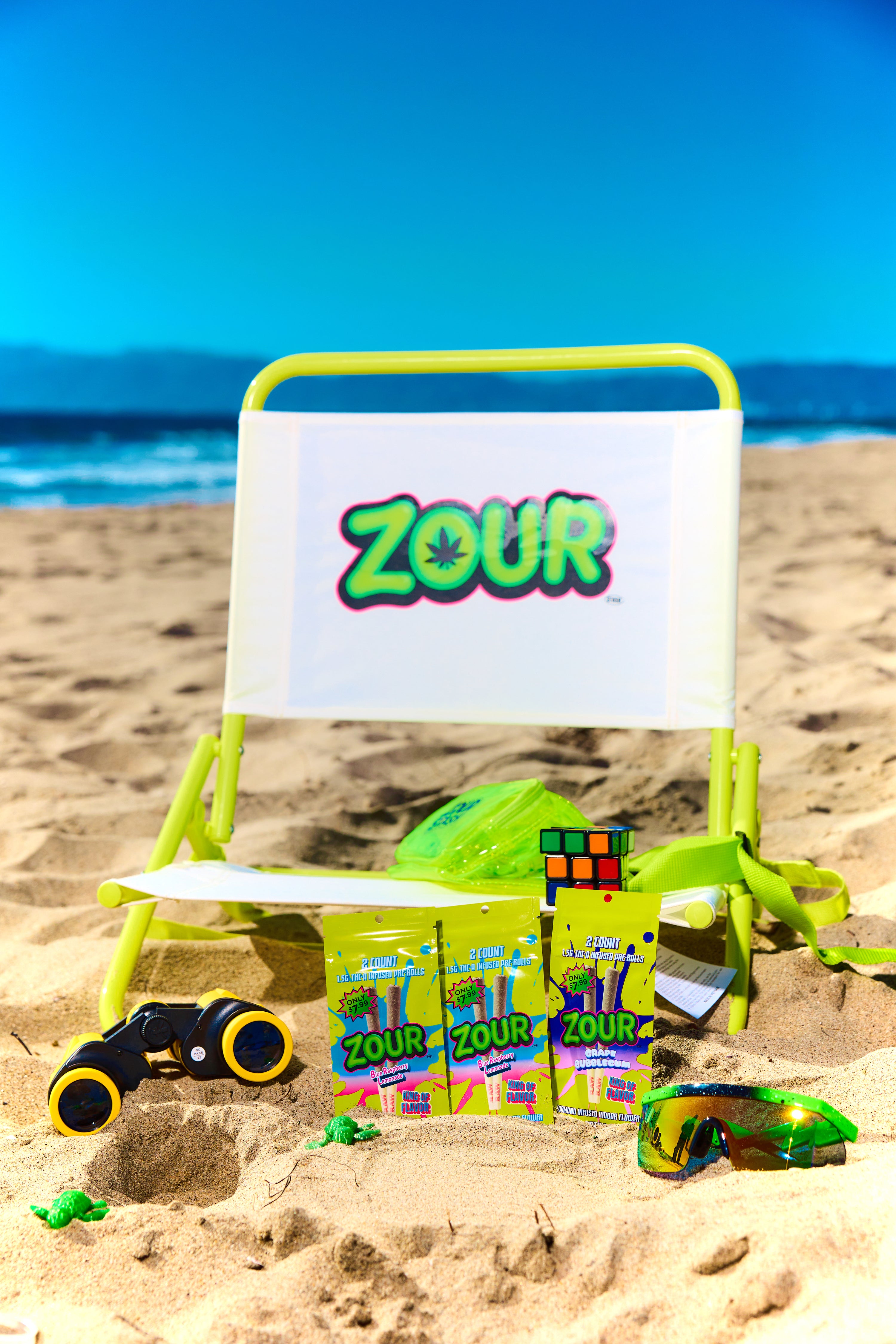 A beach-themed product display featuring a lime green folding beach chair with a white backrest printed with the ZOUR logo, set on a sandy beach with ocean and blue sky in the background. In front of the chair are several ZOUR-branded candy packages, a green bag, a small multicolor cube, binoculars, and sunglasses.