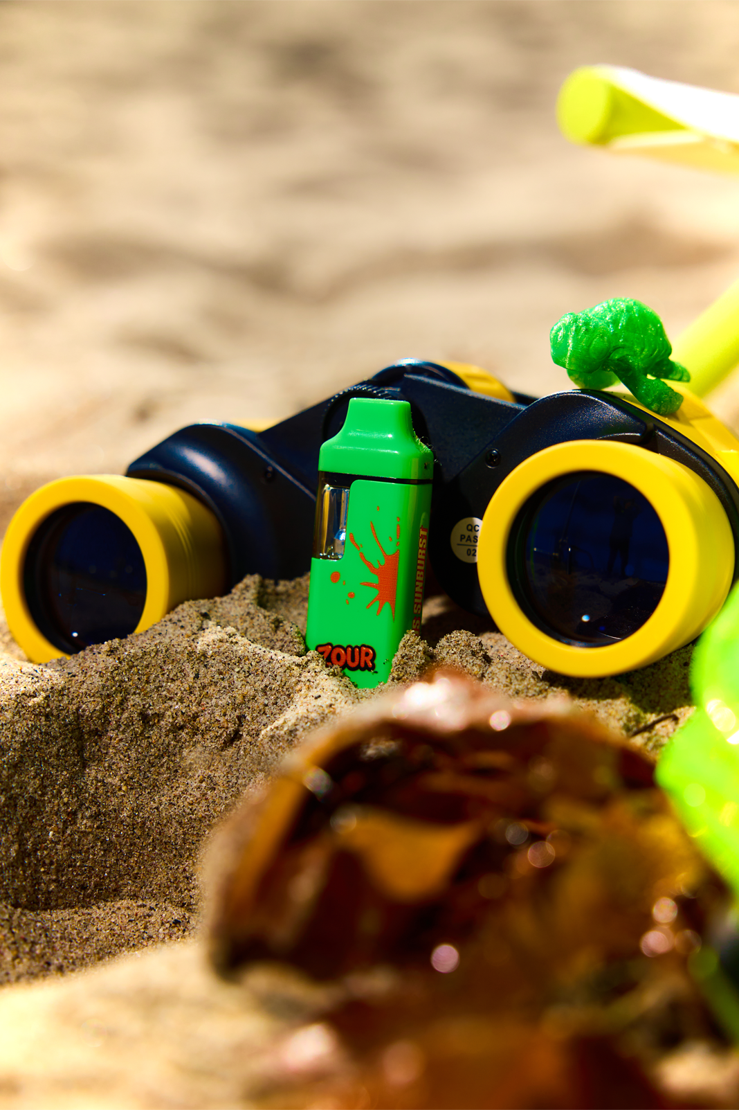 A close-up beach scene showing a pair of yellow and black binoculars resting in sand, with a small green vape labeled 'ZOUR' placed in front of them.